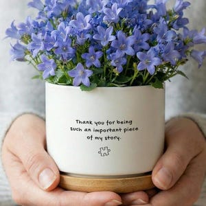 May include: A white ceramic planter filled with vibrant blue flowers. The planter has a wooden base and features the text "Thank you for being such an important piece of my story." A puzzle piece graphic is below the text.