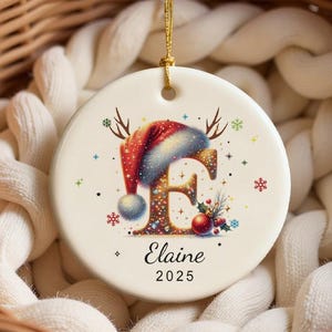 Puede incluir: Un adorno navideño redondo con una anilla dorada para colgar. El adorno presenta un diseño festivo con un gorro de Papá Noel, cuernos y la letra "F". El nombre "Elaine" y el año "2025" están impresos debajo.