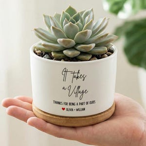 Puede incluir: Una maceta de cerámica blanca con una planta suculenta, sobre una base de madera. La maceta tiene el texto "It takes a Village" y "Thanks for being a part of ours" con nombres debajo. La suculenta tiene hojas verdes y verde claro.