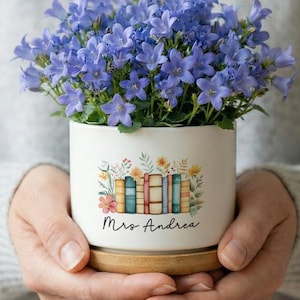 Może przedstawiać: Biała ceramiczna doniczka z kwiatowym i książkowym wzorem, wypełniona niebieskimi kwiatami. Doniczka stoi na drewnianej podstawie i jest spersonalizowana imieniem "Mrs Andrea".