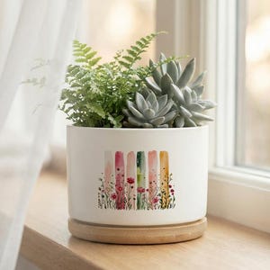 May include: A white ceramic planter with a watercolor design of colorful vertical stripes and small flowers. The planter holds a variety of green plants, including succulents and ferns, and sits on a wooden saucer.