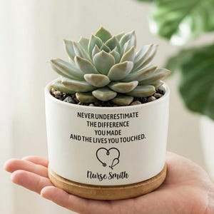 Może przedstawiać: Biała ceramiczna doniczka z sukulentem, na drewnianej podstawie. Doniczka zawiera napis "NEVER UNDERESTIMATE THE DIFFERENCE YOU MADE AND THE LIVES YOU TOUCHED." z sercem i stetoskopem oraz imieniem "Nurse Smith."