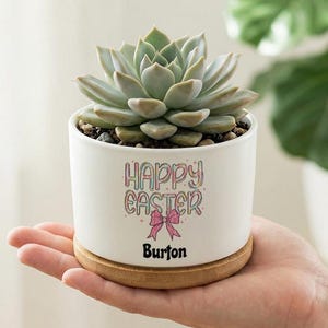 May include: A white ceramic planter with a succulent plant. The planter has "HAPPY EASTER" text, a pink bow, and the name "Burton". The planter sits on a round wooden base.