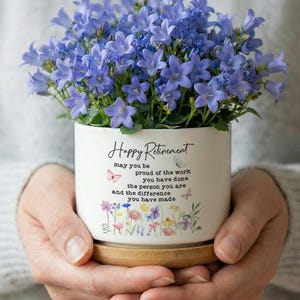 Könnte beinhalten: Ein weißer Keramiktopf mit der Aufschrift "Happy Retirement" und einem Blumendesign, gefüllt mit leuchtend blauen Blumen. Der Topf steht auf einer Holzuntertasse und wird von einer Person gehalten.