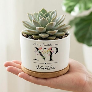 May include: A white ceramic planter with a succulent plant, resting on a bamboo coaster. The planter has the text "Nurse Practitioner" above the letters "NP" with floral accents, and the name "Martha" below. The succulent has pale green leaves.
