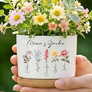 Op de afbeelding: Een witte keramische bloempot met de tekst "Mama's Garden" en bloemenillustraties, gevuld met kleurrijke bloemen. De pot staat op een houten schotel. De bloemen omvatten roze, gele en witte bloemen.