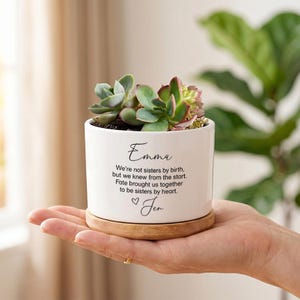 May include: White ceramic planter with a succulent plant, resting on a wooden saucer. The planter has the name "Emma" and a quote about sisterhood. The text is in black script. A heart symbol and the name "Jen" are also included.