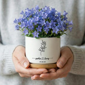 Könnte beinhalten: Ein weißer Keramik-Blumentopf gefüllt mit leuchtend blauen Blumen, der auf einer Holzuntertasse steht. Der Topf hat eine schwarze botanische Illustration und die Namen "Sophia & Lucas" mit einem Herzsymbol und dem Datum "10.26.25".
