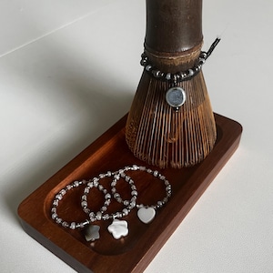 May include: A dark wooden tray holds three beaded bracelets with charms, including a star, flower, and heart. A dark brown bamboo whisk and a bracelet with a circular charm are also on the tray. The bracelets are silver and white.