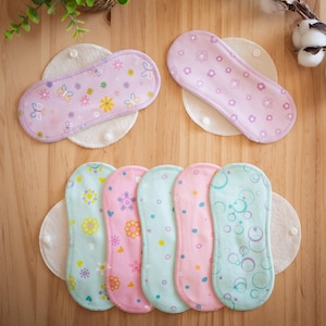 May include: An assortment of reusable cloth menstrual pads in various pastel colors, including pink, blue, and green, adorned with floral and butterfly patterns. The pads are arranged on a wooden surface, with a sprig of greenery and cotton stems in the background.
