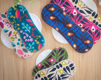 May include: An assortment of reusable cloth menstrual pads with colorful floral and leaf patterns. The pads are in various colors, including teal, pink, and navy, and rest on white circular pads. The image is taken on a light wooden surface.