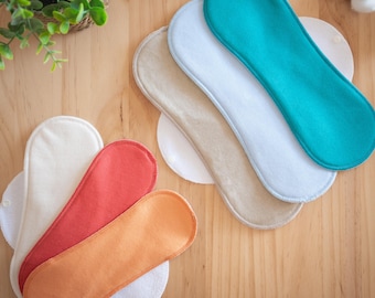 May include: An assortment of reusable cloth menstrual pads in various colors, including teal, white, beige, coral, and orange. The pads are arranged on a wooden surface, with a small plant and cotton sprigs in the background.