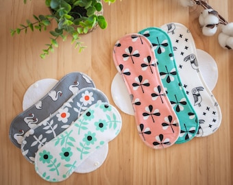 May include: Reusable cloth menstrual pads in various patterns. Designs include swans, flowers, and abstract shapes in colors like gray, pink, teal, and white. The pads are arranged on a wooden surface.