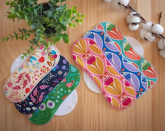 May include: A collection of reusable cloth menstrual pads in various floral patterns. The pads feature designs with cherries, flowers, and leaves in shades of pink, blue, green, and white. They are arranged on a wooden surface with a small plant and cotton branch.