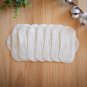May include: A stack of seven off-white, reusable cloth menstrual pads with rounded edges and a button closure. The pads are arranged on a light brown wooden surface, with a sprig of cotton and greenery in the background.