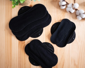 May include: Several black reusable cloth menstrual pads are arranged on a light wood surface. The pads are oval-shaped with a snap closure. Cotton sprigs and a small plant in a woven basket are in the background.