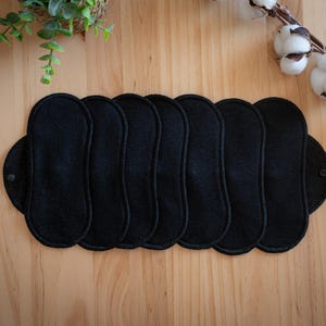 May include: A collection of seven black reusable cloth menstrual pads arranged on a wooden surface. Each pad has a rounded shape with a button closure. The pads are made of a soft, absorbent material. Decorative greenery and cotton branches are in the background.