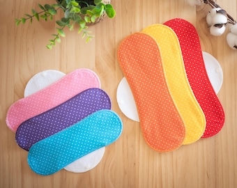 May include: A collection of reusable menstrual pads in various colors, including pink, purple, and teal, each with a white polka dot pattern. Additional pads in orange, yellow, and red are also visible. The pads are arranged on a wooden surface.
