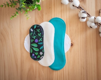 May include: Three reusable menstrual pads in different colors and patterns are displayed on a wooden surface. One pad is black with a floral design, another is white, and the third is teal. Cotton sprigs and greenery are in the background.