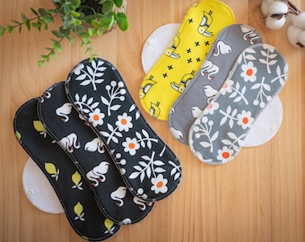 May include: A collection of reusable cloth menstrual pads in various patterns. Designs include white flowers with orange centers, swans, and lemons on black, gray, and yellow backgrounds. Each pad is placed on a white circular pad.