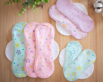 May include: An assortment of reusable cloth menstrual pads in various pastel colors, including pink, light blue, and lavender. Each pad features a floral or butterfly pattern and is placed on a white, circular backing. The pads are arranged on a wooden surface.
