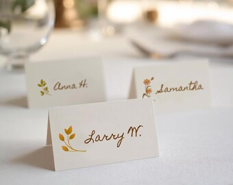 Puede incluir: Tres tarjetas plegadas con nombres escritos en escritura marrón. Cada tarjeta tiene un pequeño diseño floral o de hojas en dorado. Las tarjetas están sobre un mantel blanco, con copas y otros cubiertos en el fondo.