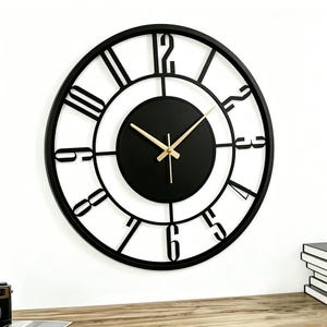 May include: A large circular wall clock featuring a black metal frame with open-work cutouts for the numbers one through twelve. The center consists of a solid black circular dial with thin gold-colored hour, minute, and second hands.