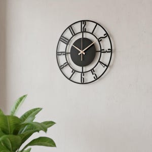 May include: A black, round wall clock with a minimalist design. The clock features a black face with light-colored hands and a black, open-faced frame with Roman numerals. The clock is mounted on a white wall.