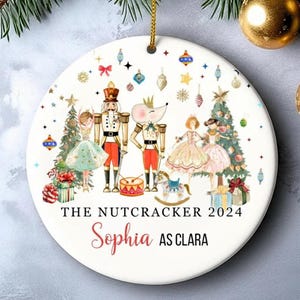 Könnte beinhalten: Runder, weißer Keramik-Weihnachtsschmuck mit Nussknacker-Design. Die Illustration zeigt Figuren, Weihnachtsbäume und Geschenke. Der Text auf dem Ornament lautet "THE NUTCRACKER 2024" und "Sophia AS CLARA". Ein goldener Faden ist zum Aufhängen befestigt.