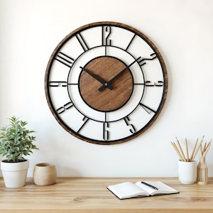 Puede incluir: Un reloj de pared grande y redondo con una esfera blanca, números romanos negros y un centro de madera marrón oscuro. El reloj tiene manecillas negras y un marco de madera marrón oscuro. El reloj está montado en una pared blanca.