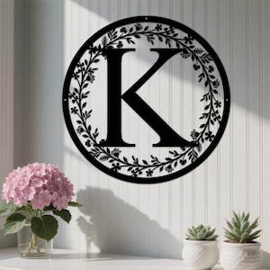 May include: Black metal monogram letter "K" encircled by a floral wreath. The letter is centrally positioned within a circular frame. The wall behind the monogram is white with vertical panelling. A pink hydrangea and two small succulents are in the foreground.