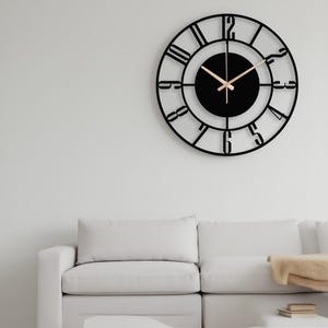 Pode incluir: Um relógio de parede preto e redondo com algarismos romanos e ponteiros dourados. O relógio está montado numa parede branca acima de um sofá branco. O design do relógio é minimalista e moderno.
