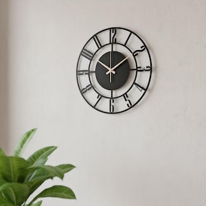 Puede incluir: Un reloj de pared negro y redondo con un diseño minimalista. El reloj presenta una esfera negra con manecillas claras y un marco negro calado con números romanos. El reloj está montado en una pared blanca.