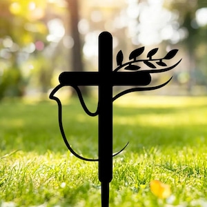 May include: Black metal garden stake featuring a cross design with a dove and olive branch silhouette. The stake is designed to be placed in the ground, adding a decorative element to outdoor spaces. The cross is approximately 30 cm tall.