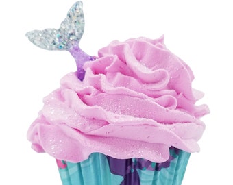 May include: A cupcake-shaped bath product with light pink, glittery whipped topping. A translucent, iridescent mermaid tail topper is inserted into the frosting. The base is a teal paper liner featuring a purple mermaid tail graphic pattern.