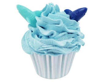 May include: A cupcake with light blue frosting and two shark-shaped decorations. The frosting is swirled and sprinkled with glitter. The cupcake sits in a white, fluted paper cup. The sharks are a light teal and dark blue.