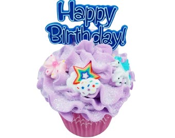 May include: A decorative cupcake with purple frosting and a blue "Happy Birthday!" topper. The cupcake is adorned with a rainbow star, a cloud with sprinkles, a flower, and a unicorn. The cupcake sits in a pink cupcake liner.