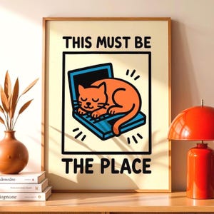 Puede incluir: Impresión artística enmarcada con un gato naranja de dibujos animados durmiendo sobre un portátil azul. El texto "THIS MUST BE THE PLACE" está impreso encima y debajo de la ilustración. La obra de arte está en un marco de madera.