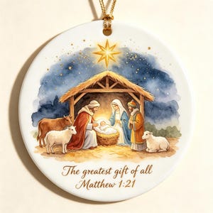 Puede incluir: Un adorno redondo de cerámica blanca con una escena de natividad en acuarela, una estrella dorada sobre un establo, figuras y animales. El texto dice: "The greatest gift of all Matthew 1:21". Incluye un cordón dorado para colgarlo.