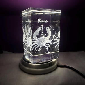Peut inclure: Un bloc de verre rectangulaire gravé au laser représentant un crabe et le mot Cancer, éclairé par une base lumineuse circulaire. Le motif inclut une constellation de points et de lignes superposée au crabe, créant un effet blanc lumineux.