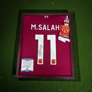 May include: Framed burgundy soccer jersey with the name "M. SALAH" and the number "11" in white. Includes a team patch and a certificate of authenticity. The jersey is displayed in a dark brown frame.