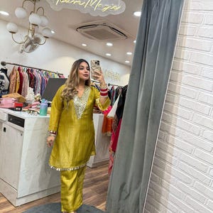 May include: A mustard yellow silk suit featuring silver embellishments on the neckline and hem. The outfit comprises a long-sleeved tunic and matching straight-leg trousers. The image also shows a mirror with the text "Darling You're Magic".
