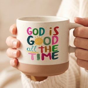 Peut inclure: Une tasse en céramique blanc cassé moucheté avec une base couleur terre cuite. L'avant présente le texte coloré "GOD IS GOOD all THE TIME" avec une petite illustration de soleil jaune intégrée dans le mot "GOOD" style peint à la main.