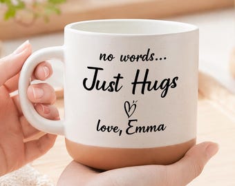 Puede incluir: Una taza de cerámica blanca con una base de color terracota natural. El frente presenta un texto impreso en negro que dice no words... Just Hugs, seguido de un pequeño icono de corazón y el nombre love, Emma en letra cursiva.