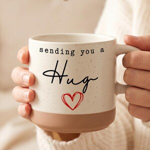 Peut inclure: Mug en céramique crème avec les mots "sending you a Hug" en écriture noire et un cœur rouge. Le bas de la tasse est de couleur marron clair. La tasse est tenue par une personne portant un pull crème.