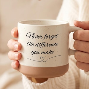 Könnte beinhalten: Cremefarbene Keramik-Tasse mit braunem Boden, mit dem Text "Never forget the difference you make" in schwarzer Schrift. Die Tasse hat eine gesprenkelte Textur und eine Herzgrafik. Ein Artikel in neutralen Farbtönen.