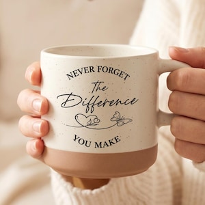 Puede incluir: Una taza de cerámica color crema con acabado moteado y una base sólida de color terracota. El frente presenta texto negro que dice NEVER FORGET The Difference YOU MAKE, acompañado de un pequeño dibujo lineal de un corazón y una mariposa.