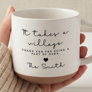 Op de afbeelding: Witte keramische mok met een gespikkelde afwerking en een bruine basis. De mok heeft de tekst "It takes a village" en "Thank you for being a part of ours." met een hartsymbool en de naam "The Smith."