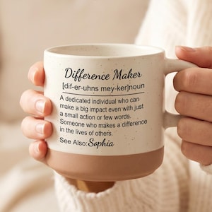May include: A beige and brown ceramic mug with the text "Difference Maker" and its definition. The mug is held by a person wearing a cream-colored sweater. The mug has a speckled finish and a rounded base.