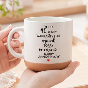 Op de afbeelding: Een witte keramische mok met een terracotta-kleurige basis met zwarte tekst: YOUR 40 year WARRANTY HAS expired SORRY no returns. HAPPY ANNIVERSARY met een klein rood hartpictogram gecentreerd onder de tekst.
